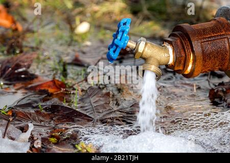 Ein kleiner Wasserhahn aus Messing im Freien. Der Hahn hat einen blauen Griff und ist an einem rostigen Rohr befestigt. Es liegt sehr nahe am Boden. Stockfoto