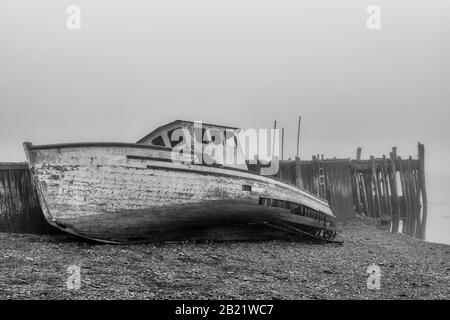 Ein Boot am Ufer neben einem alten verlassenen Kai an einem sehr nebligen Tag. Das Boot ist in sehr schlechter Form und fällt auseinander. Ebbe. Stockfoto