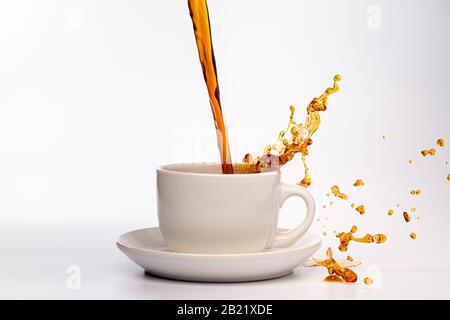 Kaffee, der in eine weiße Tasse eingießt, isoliert vor einem einfarbigen weißen Hintergrund, spritzt in alle Richtungen und erzeugt so ein Durcheinander auf der weißen Oberfläche Stockfoto