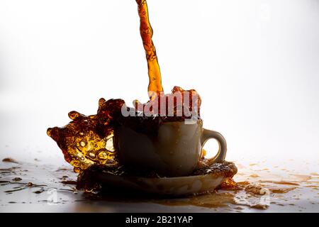 Kaffee, der in eine weiße Tasse gießt, isoliert vor einem einfarbigen weißen Hintergrund, mit Hintergrundbeleuchtung und dunklerem Kaffee, spritzt in alle Richtungen und erzeugt ein Stockfoto