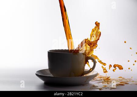 Kaffee, der in eine weiße Tasse gießt, isoliert vor einem einfarbigen weißen Hintergrund, mit Hintergrundbeleuchtung und dunklerem Kaffee, spritzt in alle Richtungen und erzeugt ein Stockfoto