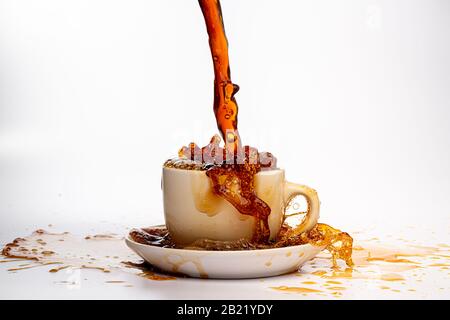 Kaffee, der in eine weiße Tasse eingießt, isoliert vor einem einfarbigen weißen Hintergrund, spritzt in alle Richtungen und erzeugt so ein Durcheinander auf der weißen Oberfläche Stockfoto