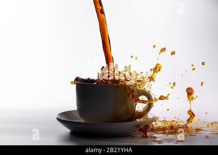 Kaffee, der in eine weiße Tasse gießt, isoliert vor einem einfarbigen weißen Hintergrund, mit Hintergrundbeleuchtung und dunklerem Kaffee, spritzt in alle Richtungen und erzeugt ein Stockfoto