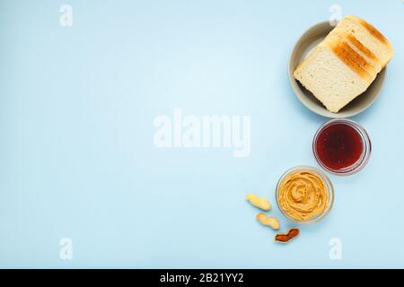 Zutaten für das kochende Frühstück mit Erdnussbutter, Brot, Toast, Marmelade. Geschälte und ungeschälte Erdnüsse, Erdnussbutter in offenem Glas in blauer Farbe Stockfoto