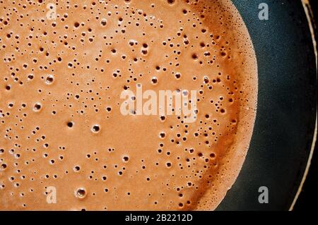 Ein Pfannkuchen aus flüssigem Schokoladen-Teig mit Blasen wird in einer schwarzen Pfanne mit Tropfen Fett und Öl auf einem Induktionsherd gebraten. Stockfoto