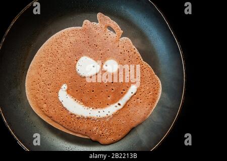 Ein Pfannkuchen aus flüssigem Schokoladen-Teig mit Blasen wird in einer schwarzen Pfanne mit Tropfen Fett und Öl auf einem Induktionsherd gebraten. Komische Küche mit Gesicht Stockfoto