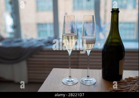 Zwei Perlmuttgläser mit Champagner, Blasen und Schaum neben einer dunklen Glasflasche gegen ein Fenster in Blautönen Stockfoto