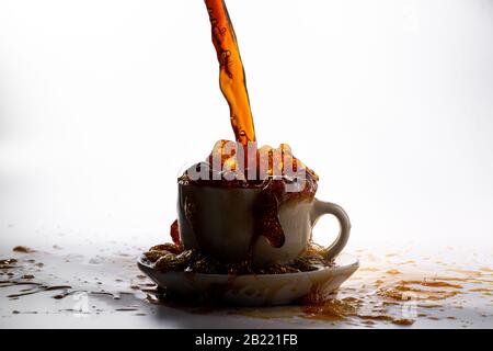 Kaffee, der in eine weiße Tasse gießt, isoliert vor einem einfarbigen weißen Hintergrund, mit Hintergrundbeleuchtung und dunklerem Kaffee, spritzt in alle Richtungen und erzeugt ein Stockfoto