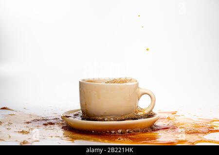 Kaffee, der in eine weiße Tasse eingießt, isoliert vor einem einfarbigen weißen Hintergrund, spritzt in alle Richtungen und erzeugt so ein Durcheinander auf der weißen Oberfläche Stockfoto