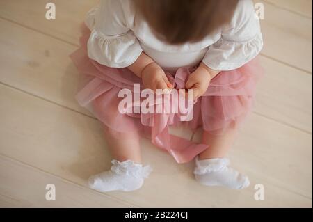 Babymädchen in einem staubigen pinkfarbenen Tüllenrock, weiße Spitzensocken mit puffigen Armen, sitzt auf einem hellen Holzboden Stockfoto