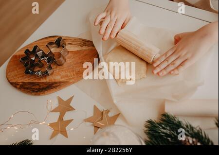 Kinderhände rollen den Teig mit einem kleinen Holzrollstift auf Backpapier, auf einem weißen Tisch Stockfoto