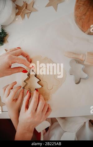 Die Hände von Mutter und Baby schneiden Plätzchen aus dem Teig mit Metallformen, auf Backpapier, auf einem weißen Tisch mit Dekor aus Papiersternen aus Stockfoto