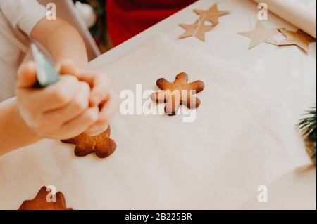 Ein Kind drückt Lebensmittelfarbstoff aus einem Röhrchen auf einen fertigen Lebkuchenplätzchen auf einem weißen Tisch Stockfoto