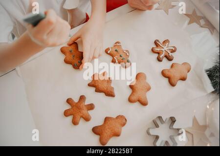 Hände malen mit einem Farbstoff aus einem Rohr auf Plätzchen, fertige Plätzchen auf einem weißen Tisch mit Backpapier Stockfoto
