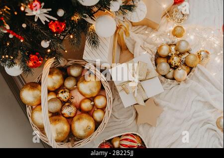 Weißes Geschenk mit goldenem Band in der Mitte riesiger Korbkorb, der oben mit roten und goldenen Kugeln gefüllt ist, um einen Weihnachtsbaum oder eine Wohnung zu schmücken, d Stockfoto