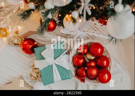 Blaues Geschenk mit einem weißen Band in der Mitte riesiger Korbkorb, der oben mit roten und goldenen Kugeln gefüllt ist, um einen Weihnachtsbaum oder eine Wohnung zu schmücken, d Stockfoto