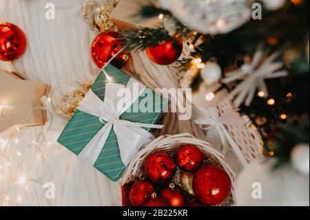 Blaues Geschenk mit einem weißen Band in der Mitte riesiger Korbkorb, der oben mit roten und goldenen Kugeln gefüllt ist, um einen Weihnachtsbaum oder eine Wohnung zu schmücken, d Stockfoto