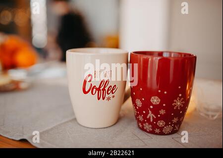 Zwei Weihnachtsbecher auf einer Baumwoll-Serviette in der Küche, ein beigefarbener Becher mit der Aufschrift über Kaffee, ein roter Becher mit Schneeflocken Stockfoto