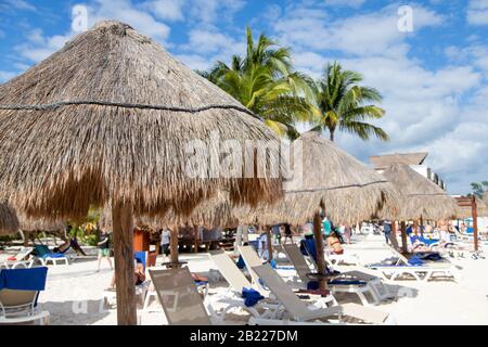 Nahaufnahme des Sonnenschirms mit Kokospalmenblatt und nicht erkennbaren Strandgängern, die sich in einer Entfernung an einem Cancun-Strand an der Riviera Maya in der Karibik entspannen Stockfoto