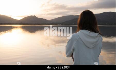 Nachdenkliche junge Frau mit langen Haaren, die von hinten gesehen wird, blickt bei Sonnenuntergang auf den See Stockfoto