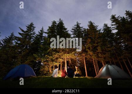 Nachtcamping in der Nähe von Lagerfeuer vor dem Hintergrund eines mächtigen Waldes. Vier Personen sitzen in der Nähe von Feuer- und Touristenzelten. Schöne Fichten unter dem Abendhimmel, auf dem die Sterne auftauchen Stockfoto