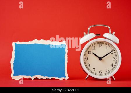 Weißer klassischer Wecker aus Vintage mit Glocken und leerem zerrissenem Papierblatt auf rotem Hintergrund. Design leer für Zeitkonzept Stockfoto