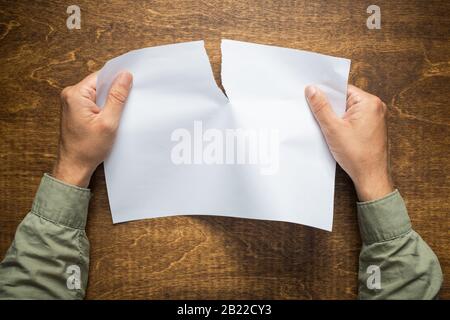 Die Hände des Mannes auf dem braunen Holztisch, der ein Stück leeres Papier reißt. Stockfoto