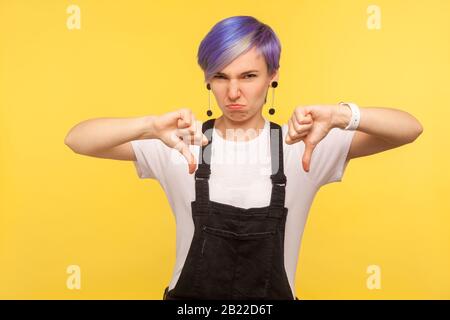 Gefällt Mir Nicht! Portrait des unzufriedenen Hüftmädchen mit violettem, kurzem Haar in Denim-Overalls, das mit Unmut und mit Daumen nach unten schaukelt, negativ di Stockfoto