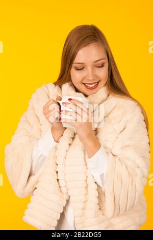 Schöne Frau in einer Pelz-Winterjacke trinkt Tee über gelbem Hintergrund - Tasse Kaffee Stockfoto