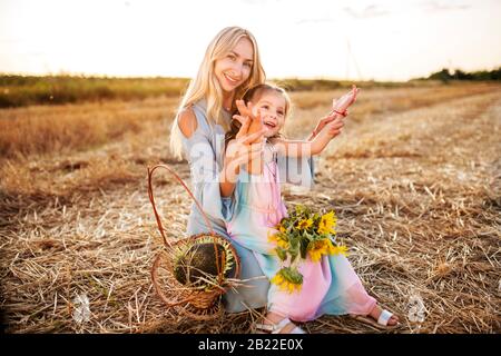 Positive junge blonde Frau spaziert und spielt mit ihrer hübschen kleinen Tochter auf einem Feld und sammelt an einem warmen Sommerabend einen Blumenstrauß mit Sonnenblumen. S Stockfoto