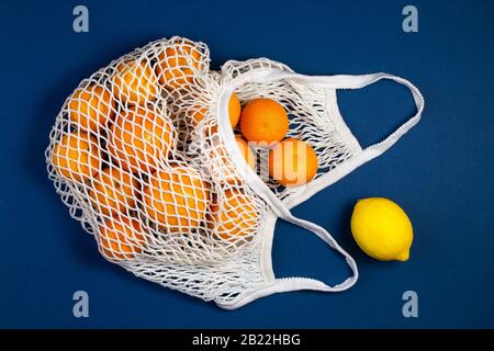Einkaufstasche aus Mesh mit Mandarinen, Zitronen auf blauem klassischen Hintergrund. Flache Lage, Draufsicht. Kein Abfall, Kunststofffreies Konzept. Gesundes, sauberes Essen stirbt Stockfoto