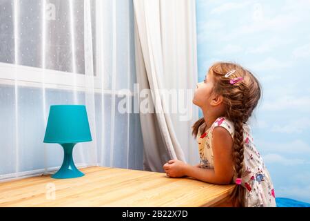 Kleines junges Mädchen, Kind auf dem Tisch angelehnt, das in der Nacht einsam im hellen Raum durch das Fenster blickt. Ich warte darauf, dass jemand zurückkommt Stockfoto