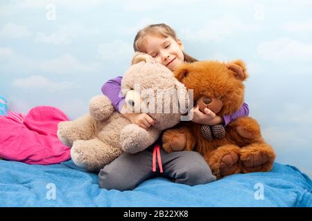 Kleines Mädchen, lächelndes Kind, das auf dem Bett sitzt und zwei große Teddybären umarmt, fröhliches Kind, das ihre beiden Spielzeuge in der Nähe hält. Blauer Raum Wolken Hintergrund, Herz Stockfoto