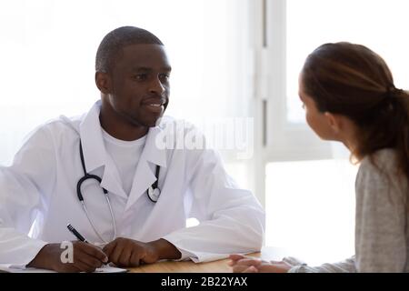 Lächelnder afroamerikanischer Arzt, der in der Klinik auf die weibliche Patientin hört Stockfoto