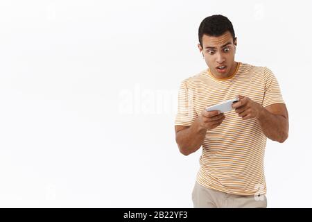 Überrumpelte und amüsante hispanische Männer in drahtlosen Kopfhörern, die aufregendes Online-Handy-Spiel spielen, Smartphone horizontal halten, Telefon neigen Stockfoto