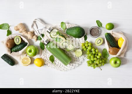Verschiedene grüne Obst- und Gemüsesorten mit Flaschen Smoothie und Wasser auf weißem Holztisch Draufsicht. Umweltfreundliches Einkaufskonzept. Detox di Stockfoto
