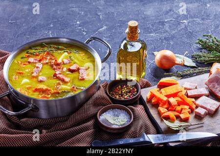 Klassische deutsche Erbsenunterlage mit Speck, Schweinehinken, Karotte, Kartoffel mit Thymian - winterherzhaftes Gericht auf einem Topf auf Betongrund, oliv Stockfoto