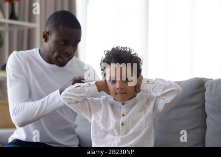 Kleiner Sohn, der Ohren bedeckt und den strengen wütenden afroamerikanischen Vater ignoriert Stockfoto