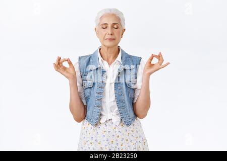 Geduldig, ruhig gesunde, moderne Großmutter mit grau gekämmtem Haar in stilvollem Denim-Hemd, Kleid, meditierendem Stand im Yoga, Mudra-Pose, geschlossenen Augen Stockfoto