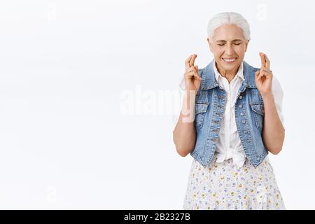 Hoffnungsvolle, aufgeregte lächelnde Seniorin hofft, dass sie wahr wird, träumt davon, erfüllen zu wollen, schließt die Augen grinsend und die Kreuzfinger Glück haben, betet Stockfoto