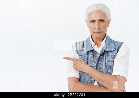 Verärgerte und verärgerte Seniorin mit grauem Haar, erfroren belästigt, Zeigefinger nach links, missfällt, wie unordentliche Zimmer sind, jemanden erschrecken, starrend Stockfoto