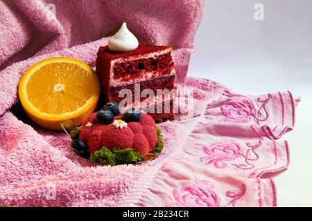 Zwei appetitliche rote Kuchen und die Hälfte saftige Orange liegen auf einem rosafarbenen Handtuch mit Fancywork Stockfoto