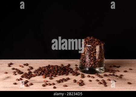 Whiskyglas voller gerösteter Kaffeebohnen und einiger auf einer Holzoberfläche verstreuter Körner Stockfoto