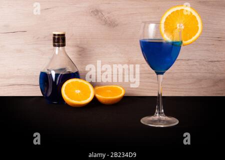 Glas mit blauem Getränk und Scheibe saftiges Orange auf der Seite, verschüttete Flasche und orangefarbene Hälften dahinter Stockfoto