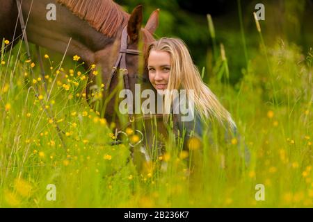 Lächelnde blonde Frau, die mit ihrem arabischen Pferd auf der Wiese sitzt Stockfoto