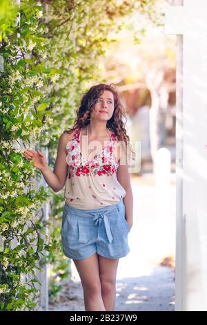 Junge Frau Mädchen, die im Sommer an einem sonnigen Tag im Garten unterwegs ist und die Blumen der Weinpflanze draußen mit Pastellmode berührt Stockfoto