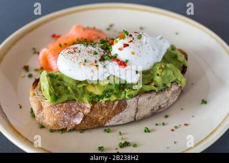 Pochierte Eier auf zertrümmtem Avocado auf Sauerteigtoast mit geräuchertem Lachs. Gesundes Frühstück mit Teller auf dem Tisch. Nahaufnahme, geringe Schärfentiefe. Stockfoto
