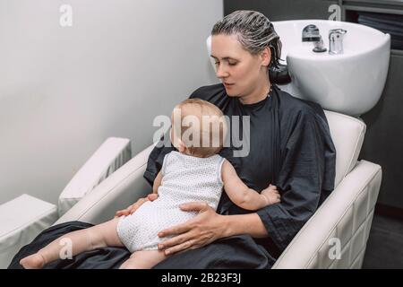 Mutter mit Baby im Schönheitssalon wartet darauf, dass die Friseursalon ihre Haare nach dem Färben waschen kann. Stockfoto