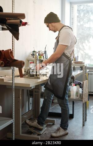 Junger Mann in Arbeitskleidung steht am Tisch mit einem Fuß auf dem Pedal, während er mit einer elektrischen Maschine Lederwaren anstellt Stockfoto