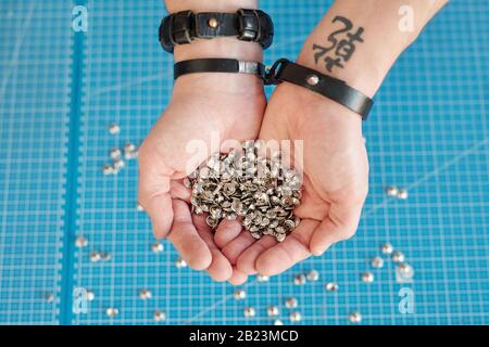 Übersicht über die Hände des Handwerkers in Lederarmbändern, die den Haufen kleiner metallischer Knöpfe über blauem Hintergrund halten Stockfoto